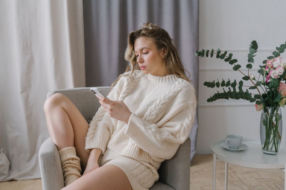 Woman in cozy sweater using smartphone in a modern indoor setting, relaxing on an armchair.