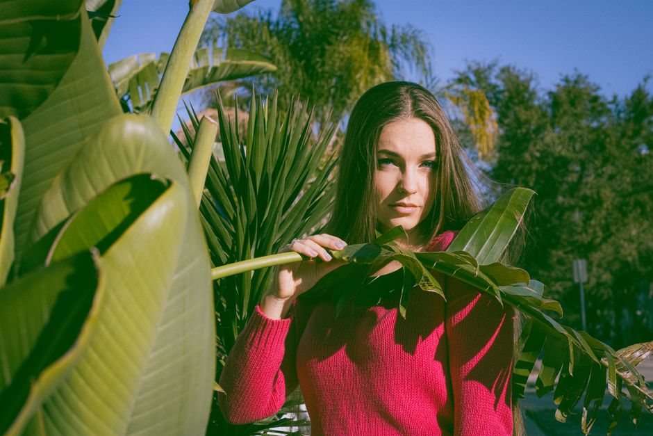 Young woman in pink sweater poses with tropical plants on a sunny day, capturing a vibrant, natural vibe.