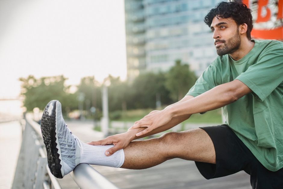 A man in a green shirt stretches his leg outdoors near a modern building, promoting a healthy lifestyle.
