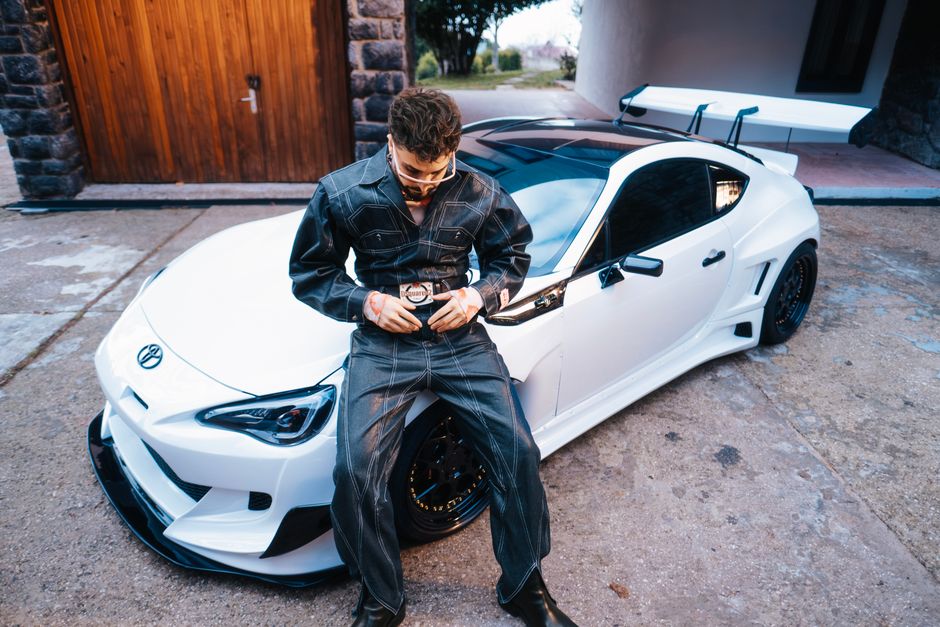 Young man in denim outfit sits on a sleek white sports car in an outdoor setting, conveying a stylish urban vibe.