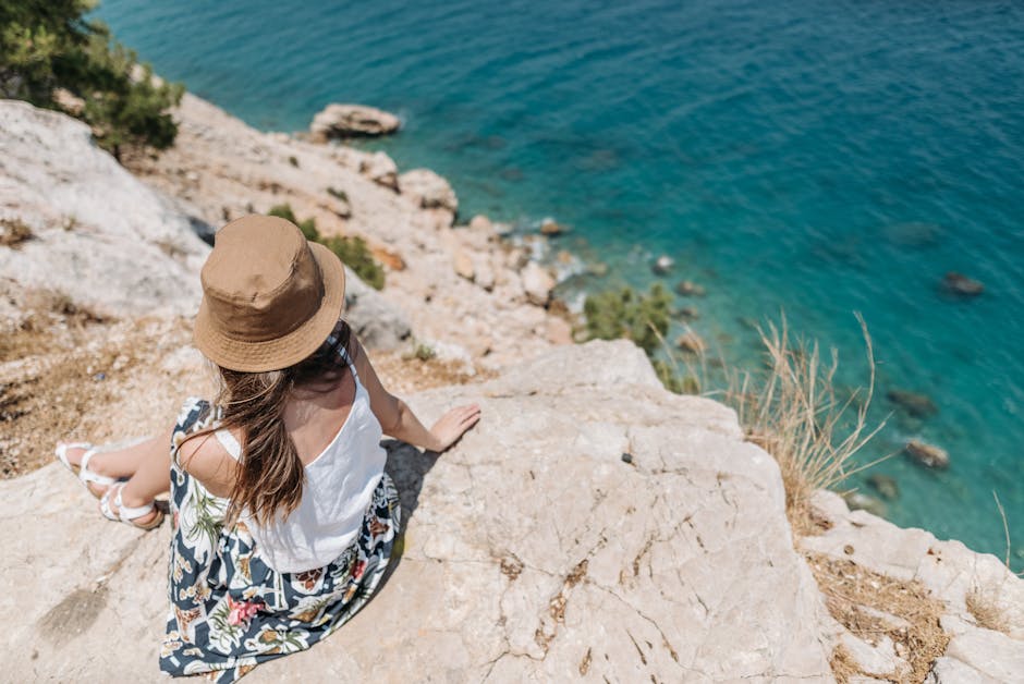 A woman sits on a rocky cliff, enjoying the summer view of turquoise waters.