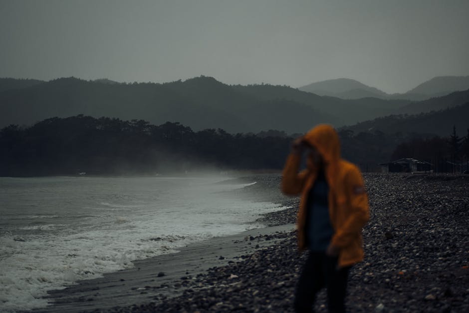 Person in yellow jacket walking on a foggy rocky beach with mist and mountains in the background.