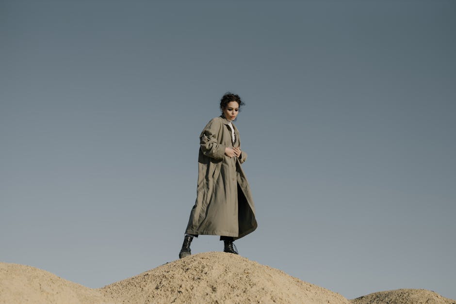 Stylish woman in trench coat stands on sandy dune under clear sky, showcasing fashion in nature.