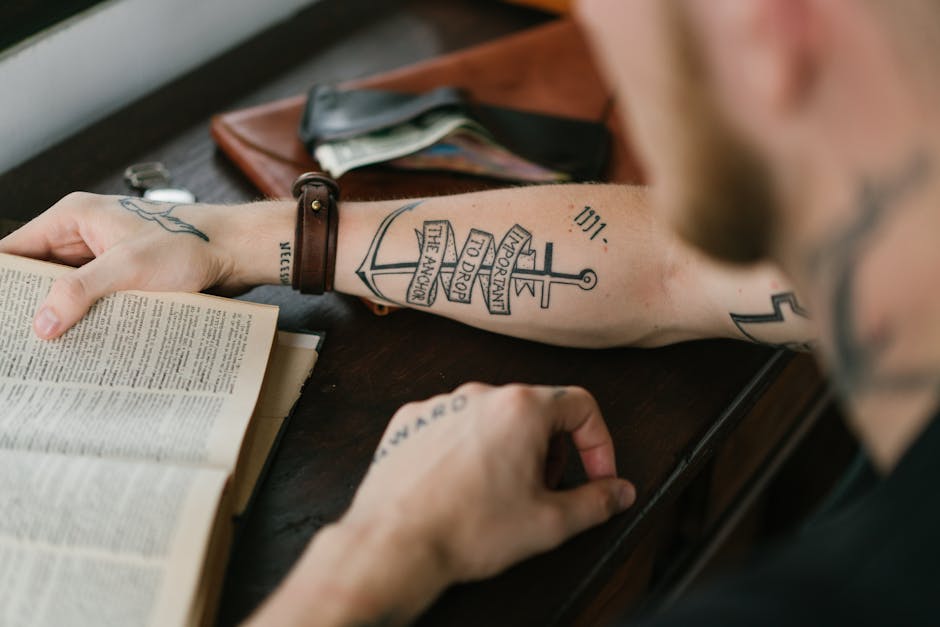 Crop masculine male with tattoo saying Important to Drop the Anchor on arm reading book while sitting at desk