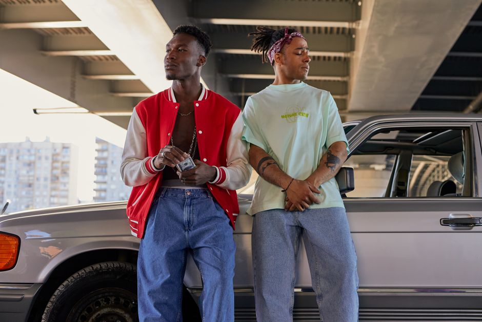 Two men leaning against a classic car under a bridge, showcasing urban fashion.