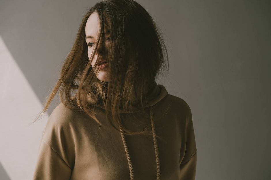 A young woman with long hair poses expressively in a hoodie, lit by soft indoor lighting.