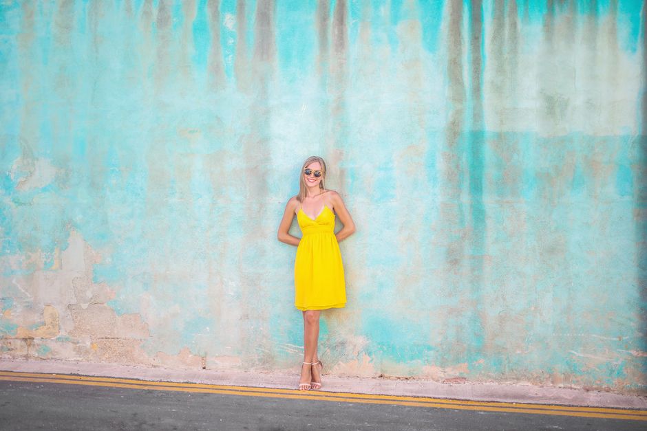 Young woman in yellow dress smiling with sunglasses against a turquoise wall outdoors.