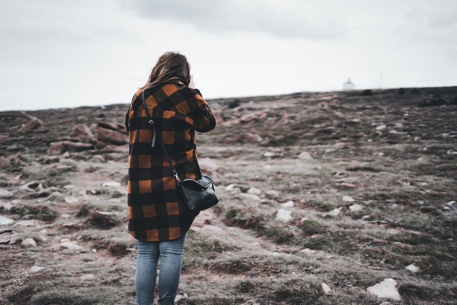 Woman in plaid coat with leather bag standing on rocky outdoor terrain.