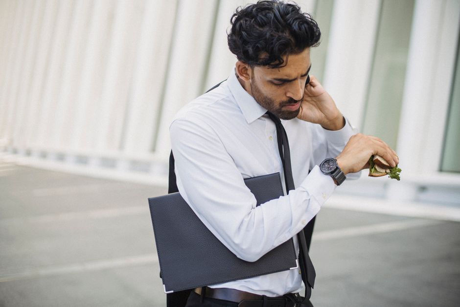 Businessman multitasking, checking time while holding a snack in an urban outdoor setting.