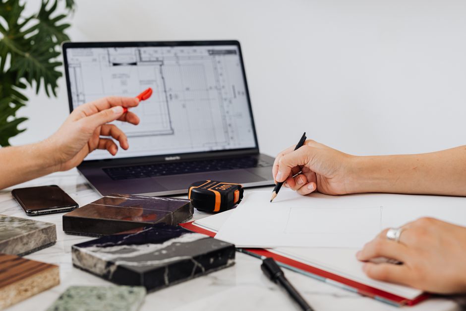 Close-up of hands sketching design plans with tile samples and a laptop, ideal for interior design contexts.