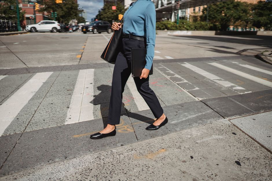 Unrecognizable female in casual wear with black bag walking on pedestrian crossing on sunny street in city with buildings on background
