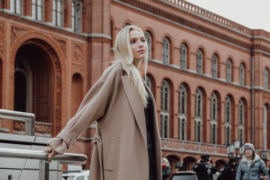 Stylish woman with long blonde hair poses elegantly in front of a historic red brick building in an urban setting.