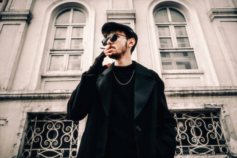 Fashionable man in black coat and hat smoking in an urban setting. Trendy and bold.