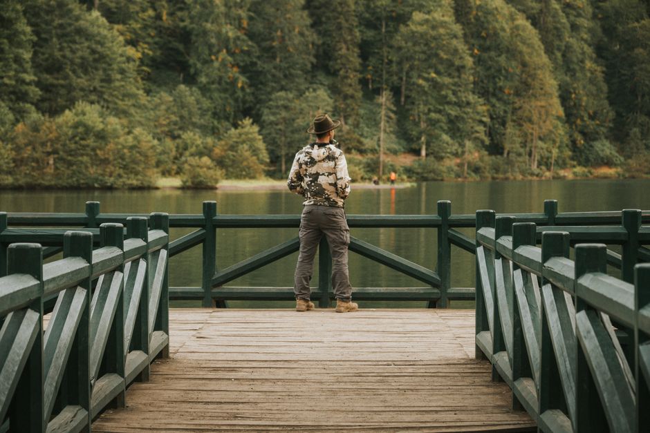 A man in a hat and jacket standing on a wooden dock, gazing at a tranquil lake surrounded by lush forest.