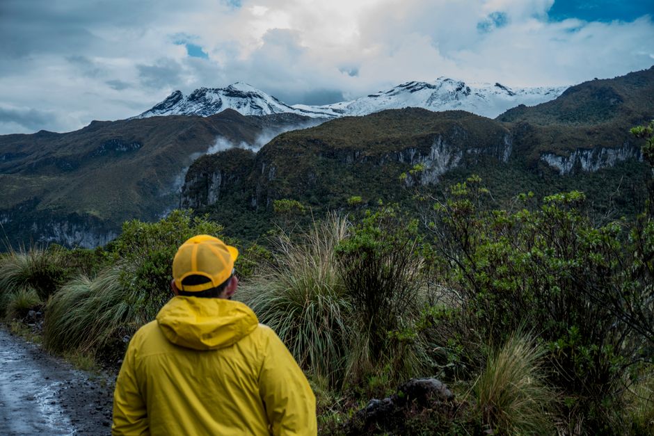 A man in a yellow jacket hikes through a scenic mountain landscape with snow-capped peaks.