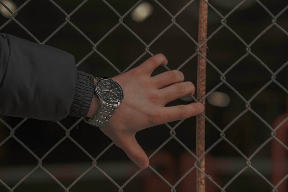 A close-up of a hand wearing a wristwatch resting on a chain link fence, captured in low light.
