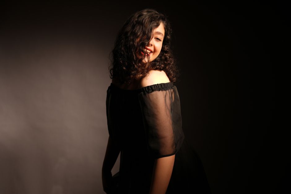 A young woman with curly hair poses confidently in soft lighting, wearing an off-shoulder dress.
