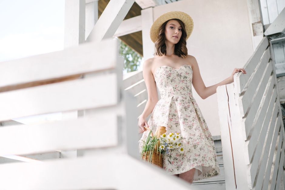 Elegant woman in a floral dress and hat holding daisies on white stairs, embracing summer style.