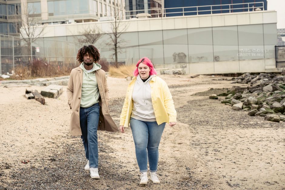 Full body of multiracial couple in jeans and outerwear strolling on embankment with stones against building during date in city
