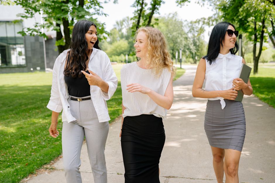 Three smiling businesswomen walk and converse in a sunny park setting, showcasing diversity and professional camaraderie.
