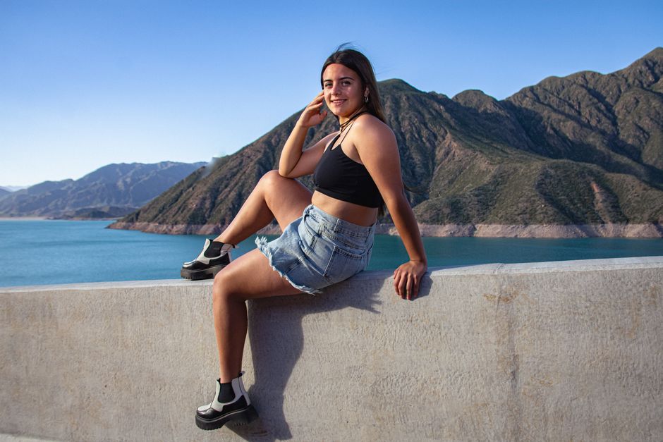 A smiling woman in casual attire sits on a ledge with a stunning mountain backdrop.
