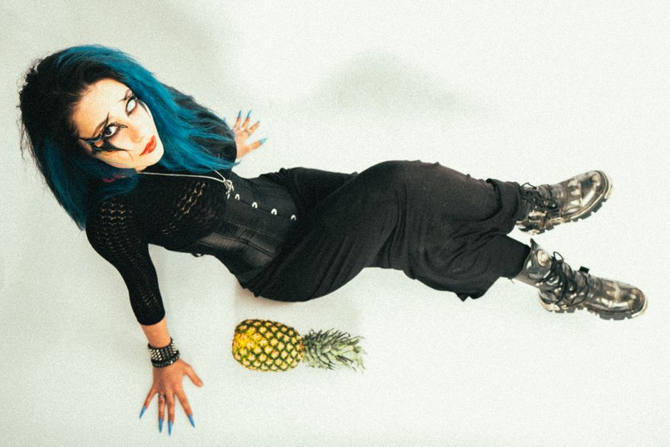 A woman with vibrant blue hair and gothic attire poses next to a pineapple on a white background.