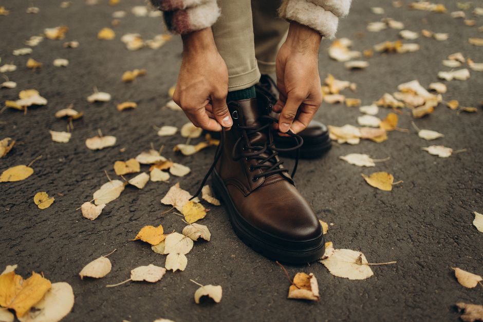 Close-up of a person tying brown leather boots on a leafy street in autumn.