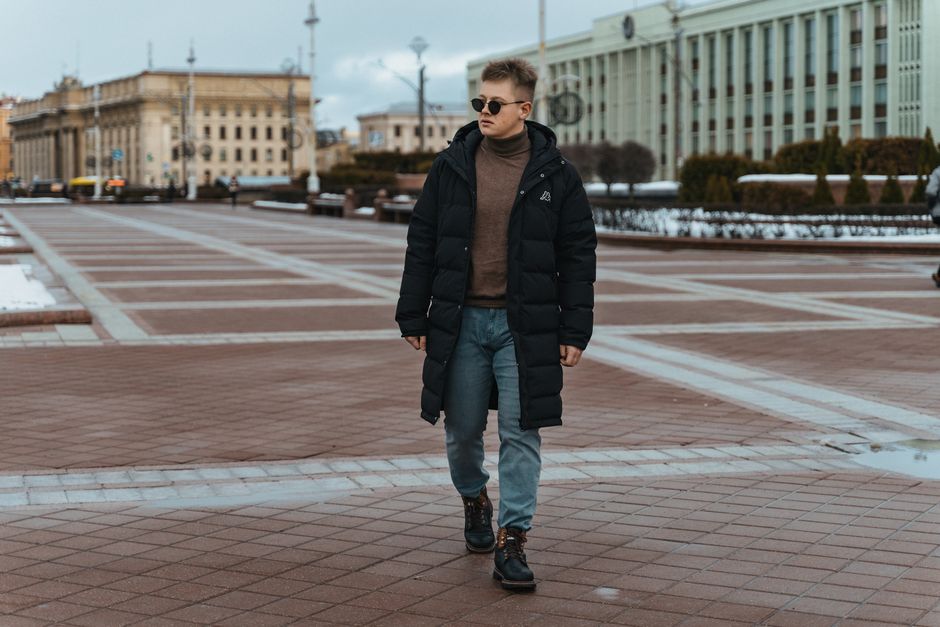 A man in a black coat walks confidently through a city square.