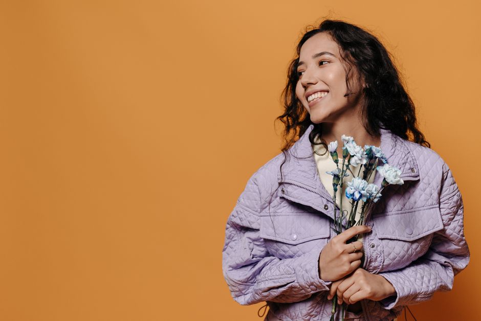 Young woman in a purple jacket smiling and holding blue carnations against a peach background.