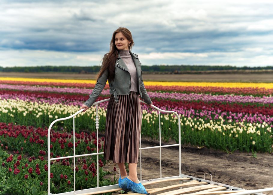 A woman stands on a bed frame surrounded by colorful tulip fields under an overcast sky in Belarus.