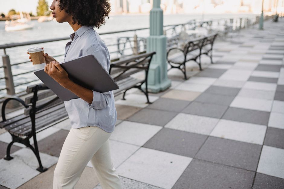 A woman strolls along a riverside promenade, holding a laptop and coffee cup.