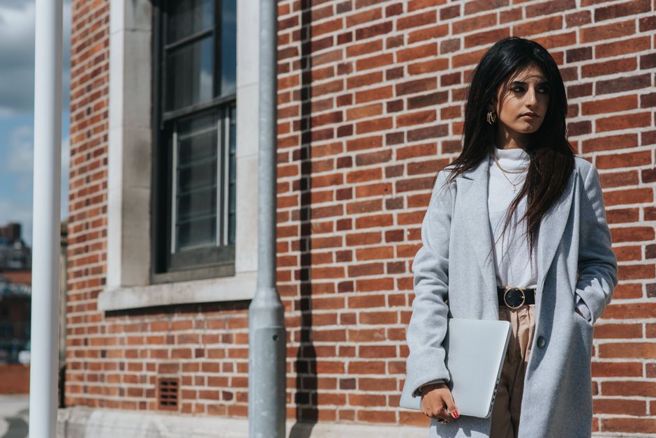 Young trendy ethnic female executive in coat standing with hand in pocket and netbook in city while looking away