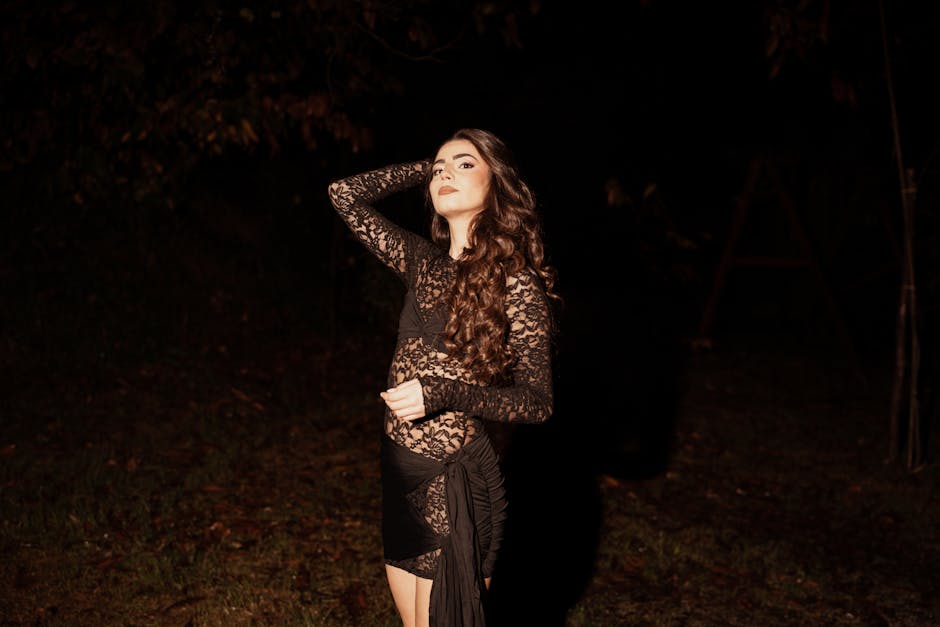 Elegant woman in a lace dress posing confidently in a dark outdoor setting, with long wavy hair.